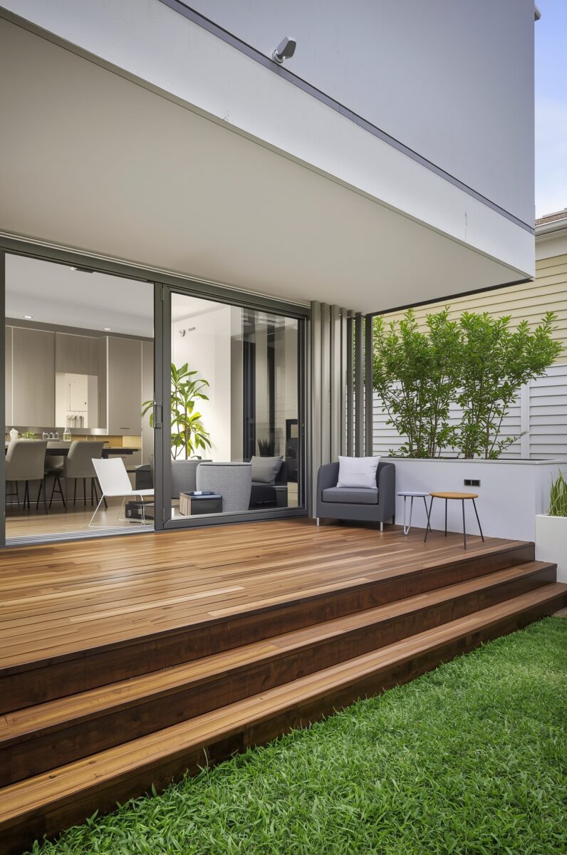Modern home outdoor timber deck with sliding glass doors, grey armchair, small side table, green lawn, and lush plants