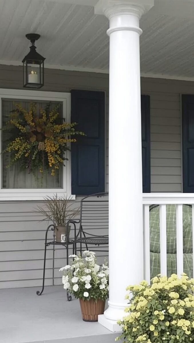 Charming front porch with white column, navy shutters, black lantern light, wrought iron chair, and white and yellow potted flowers