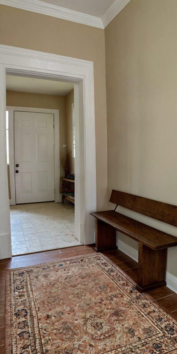 Warm beige hallway with dark wood bench, floral area rug, and white front door visible through doorway