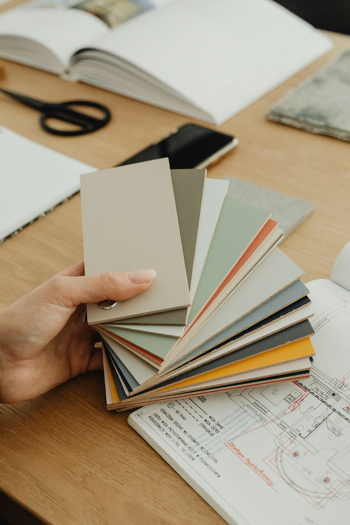 A hand fanning out paint sample cards in soft greens and neutrals over an open notebook