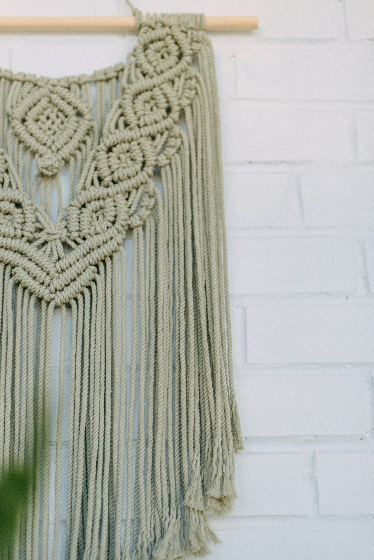 A sage green macrame wall hanging on a wooden dowel against a white brick wall