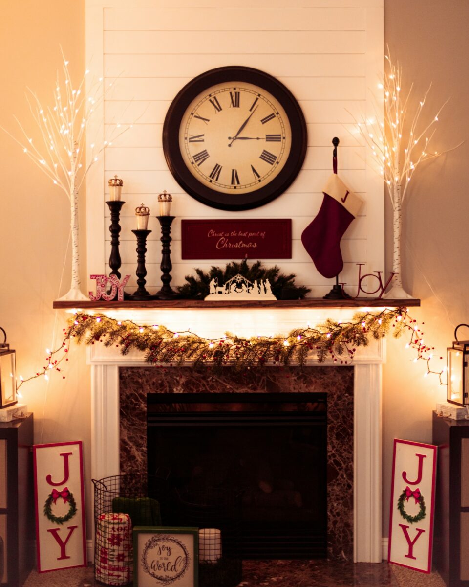 A white shiplap mantel decorated for Christmas with a large wall clock, three black candlesticks of different heights, a lit garland and a stocking