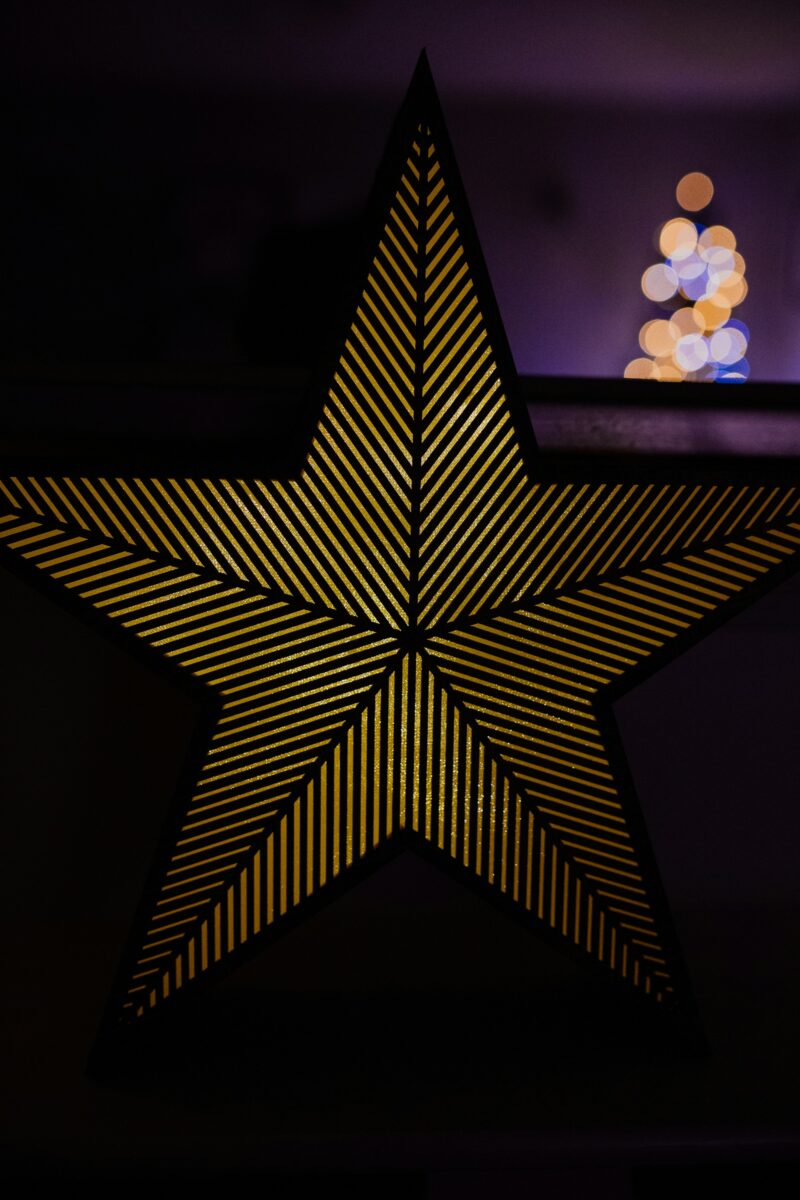 A lit star lamp with gold stripes in a dark room, a Christmas tree's lights blurred behind it