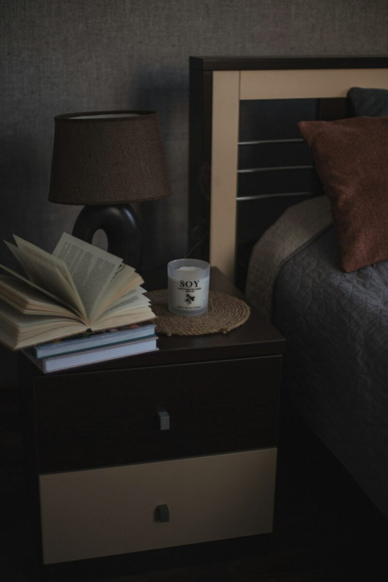 A nightstand with a lamp, an open book on a stack of books and a jar candle on a woven mat beside a bed