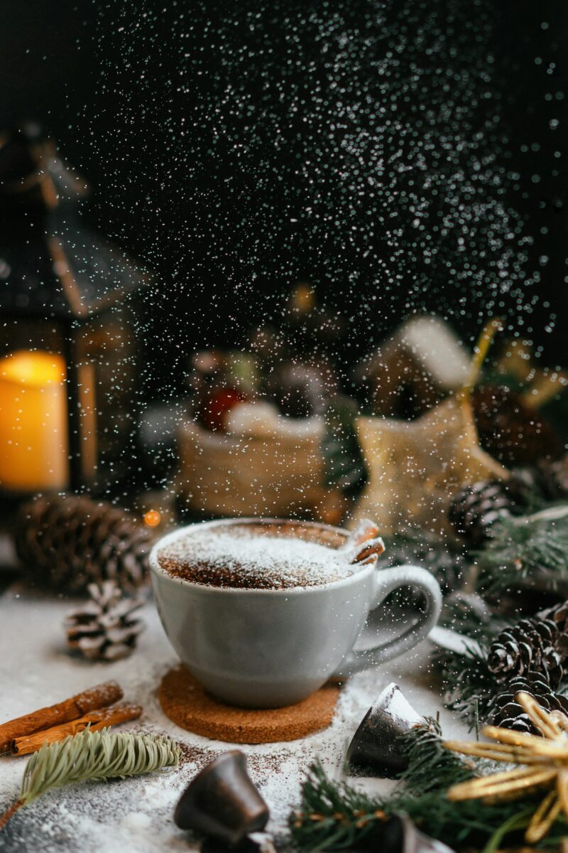 A cup of cocoa dusted with sugar on a cork coaster, pine cones, cinnamon sticks and a lantern around it