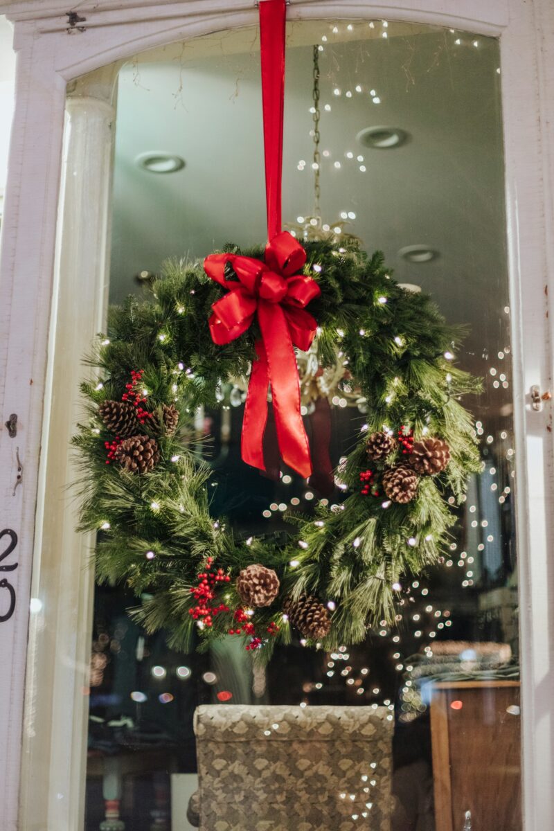 A green Christmas wreath with a red bow, pine cones and berries hung on a long red ribbon over a glass shop door