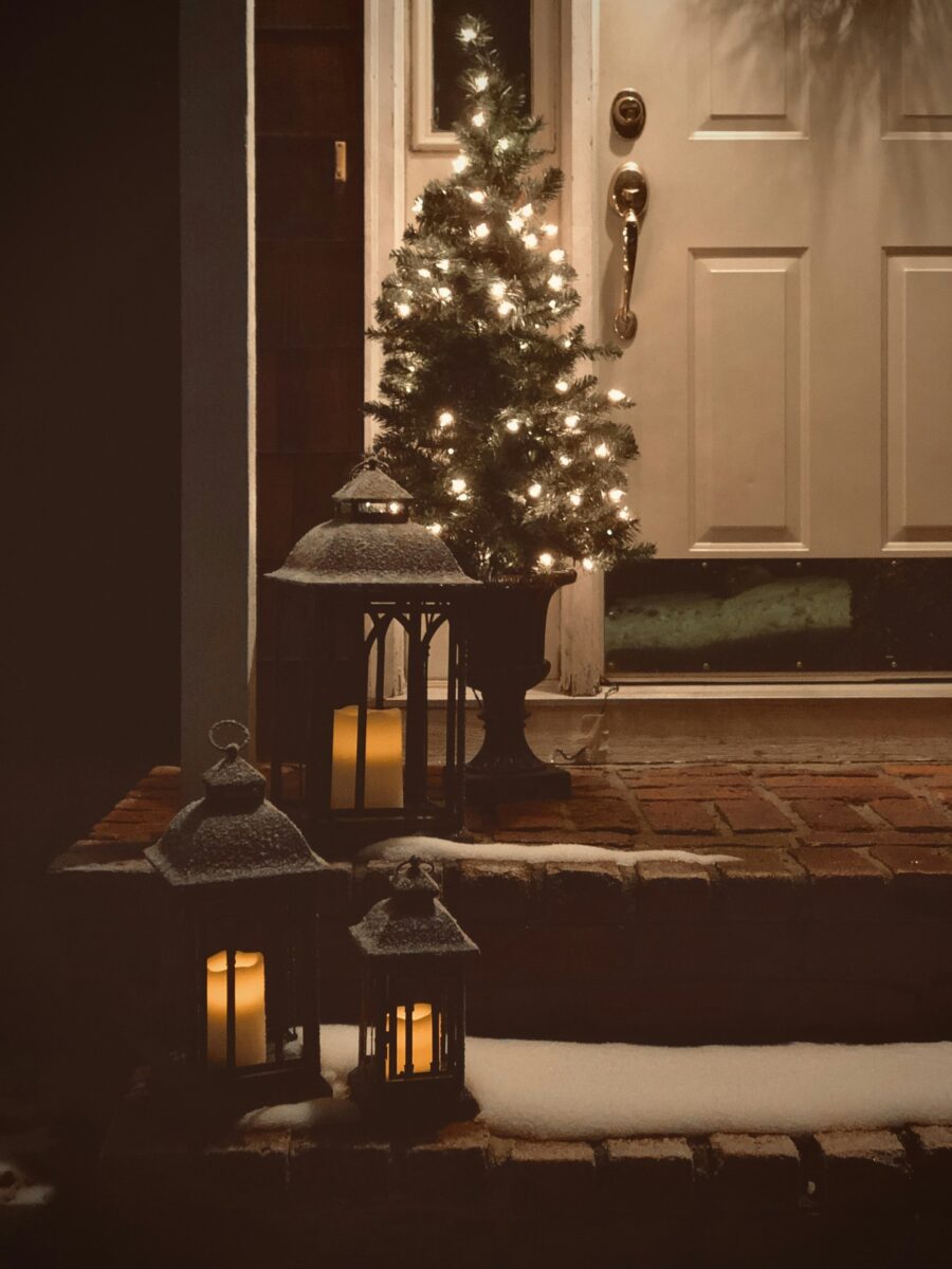 A front door at night with a small lit Christmas tree in an urn and three glowing lanterns on snowy brick steps