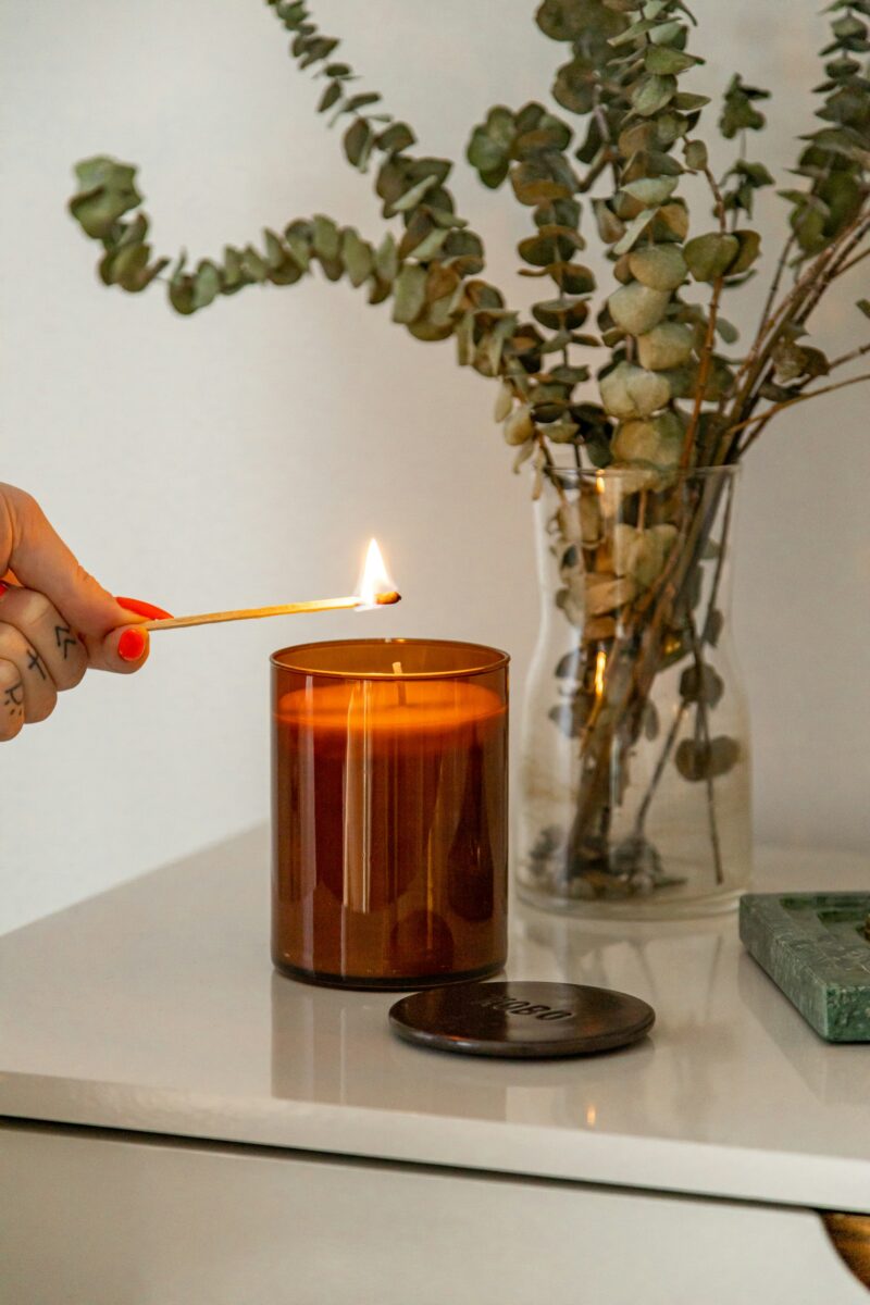 A hand lighting a candle in an amber glass jar with a match, a vase of eucalyptus behind it