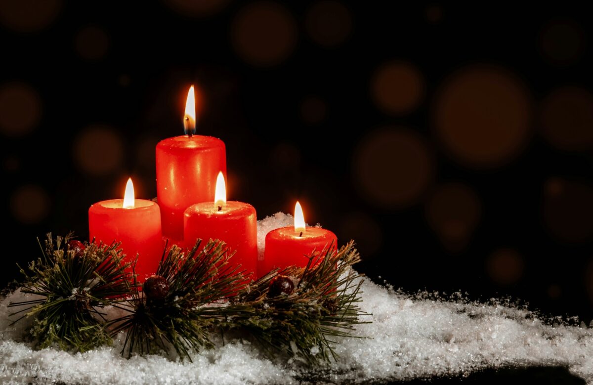 Four lit red pillar candles surrounded by pine sprigs and white snow against a dark background