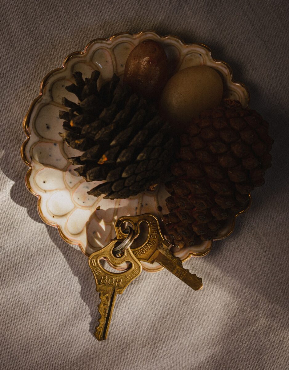 A small scalloped ceramic dish holding two brass keys, two pine cones and two smooth round stones on a linen cloth