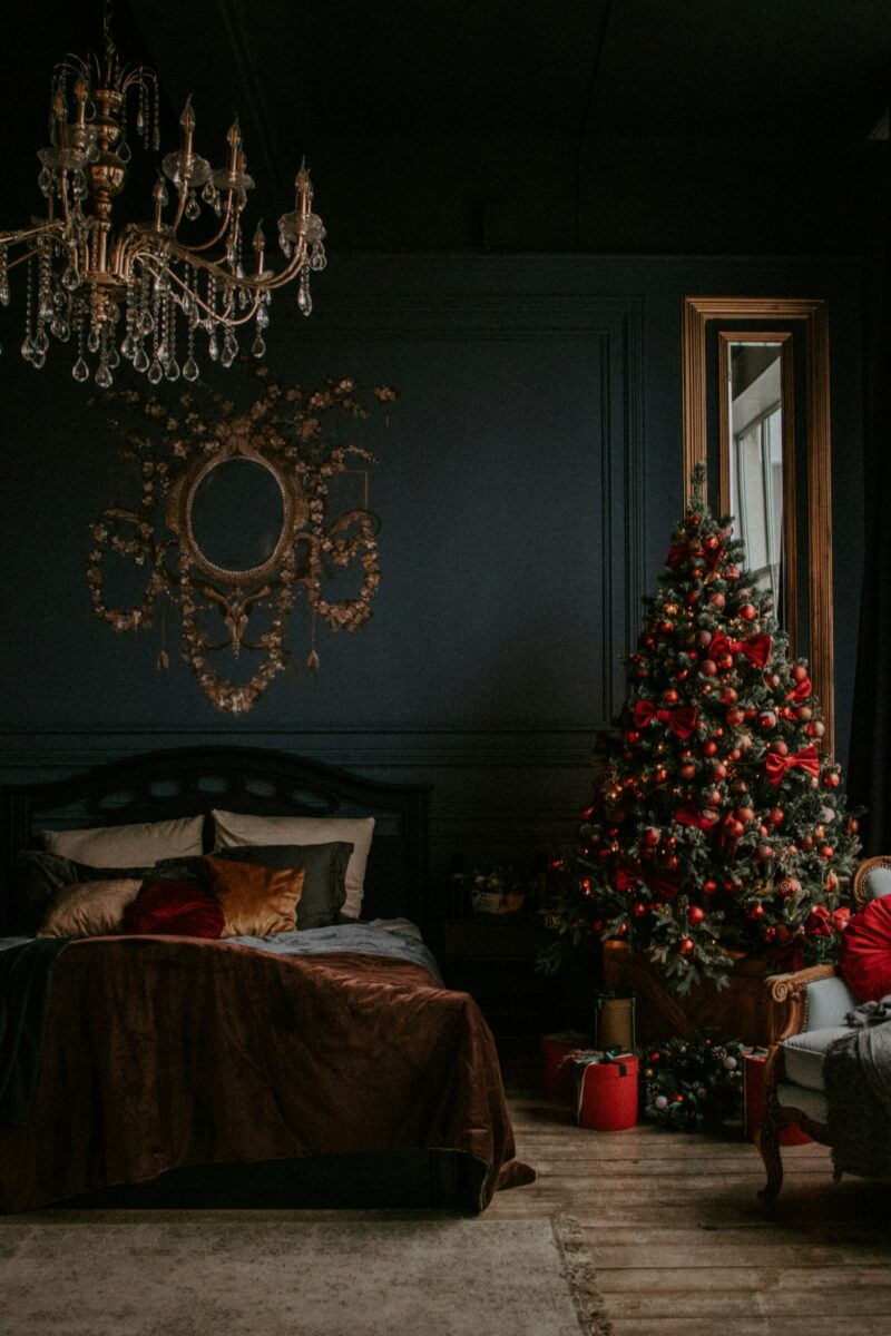 A dark green bedroom with a Christmas tree in red ornaments and bows in the corner, a velvet throw on the bed and wrapped gifts on the floor