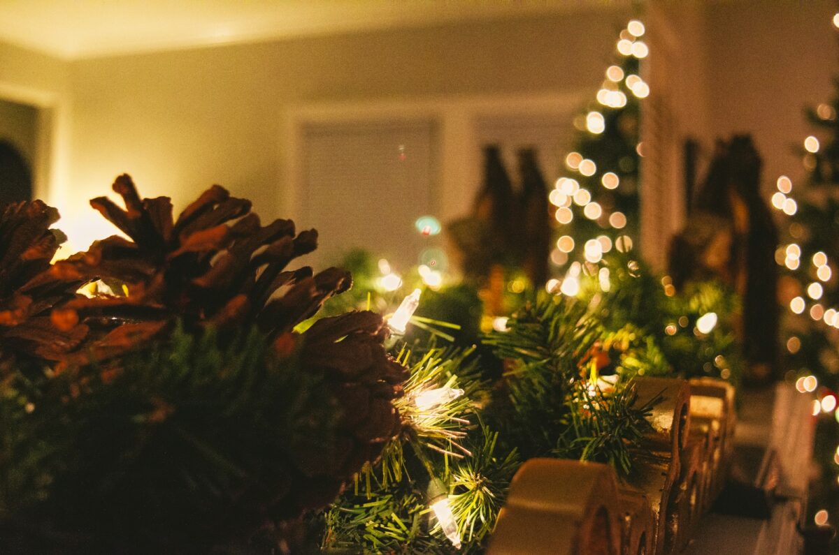 A pine garland with large pine cones and warm string lights laid along a ledge, with lit trees blurred behind