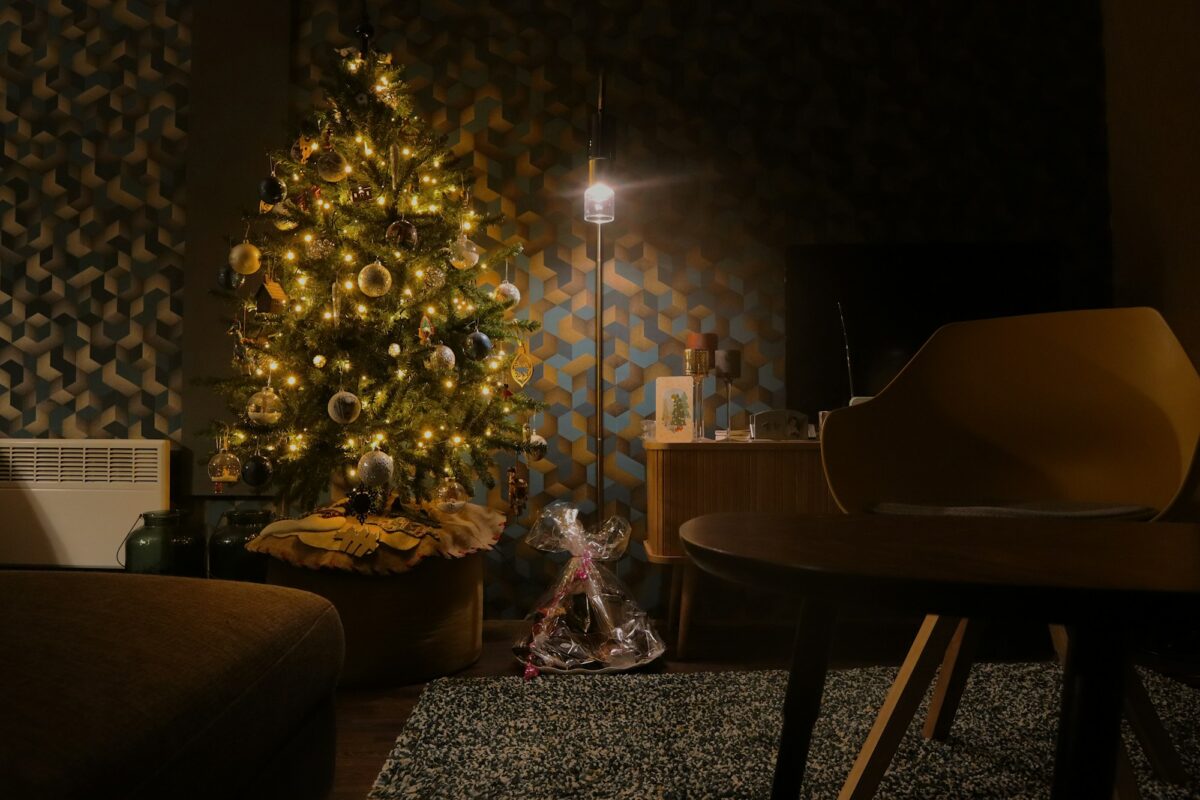 A glowing Christmas tree in a dark living room with a patterned wall, a floor lamp and armchairs