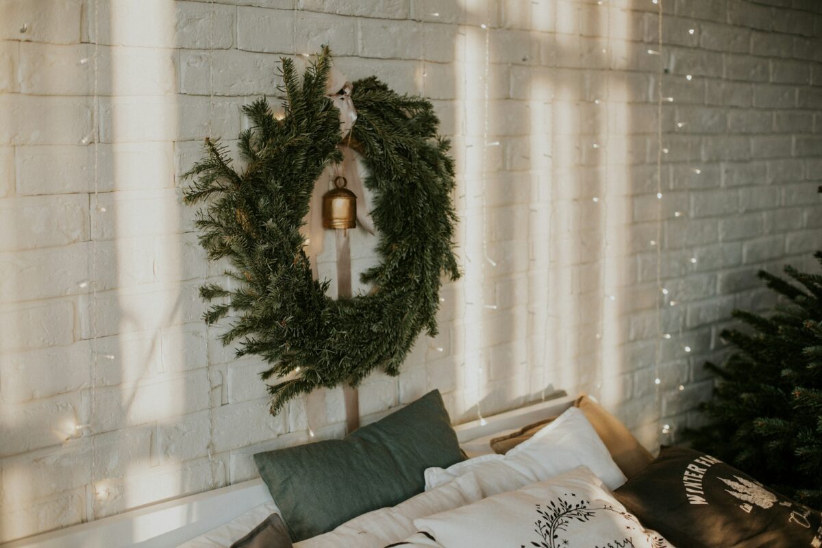 A fir wreath with a brass bell hung on a white brick wall above a bed with green and white pillows