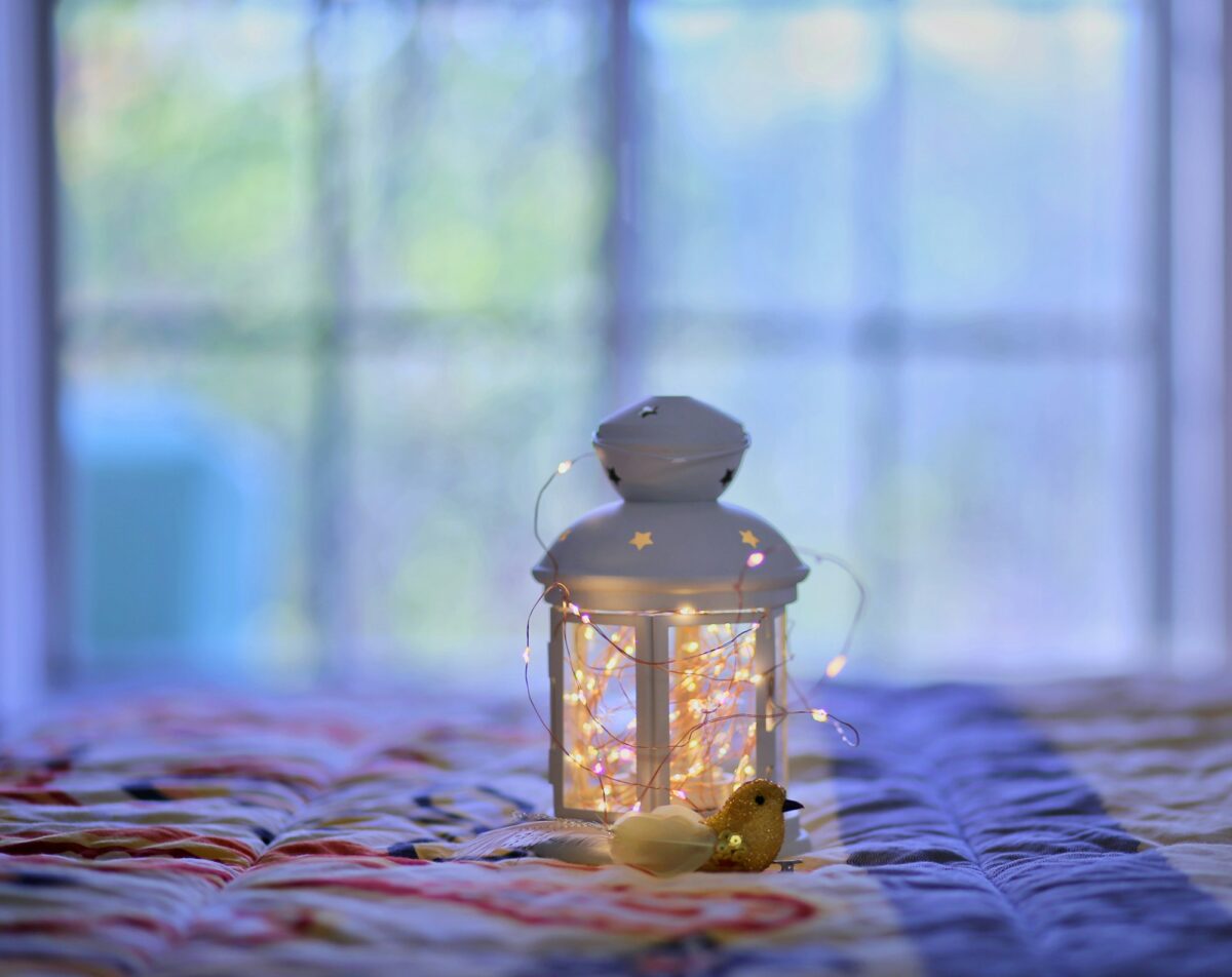 A white lantern filled with warm fairy lights sitting on a quilt in front of a window