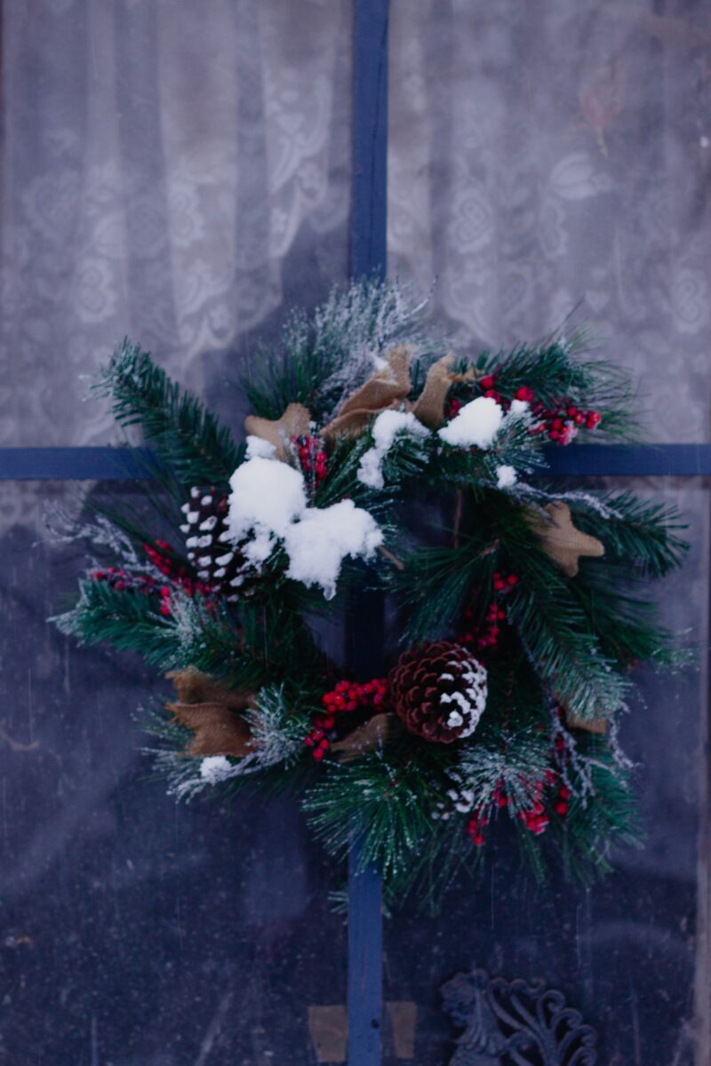 A frosted pine wreath with pine cones and red berries hung in a window in front of a lace curtain