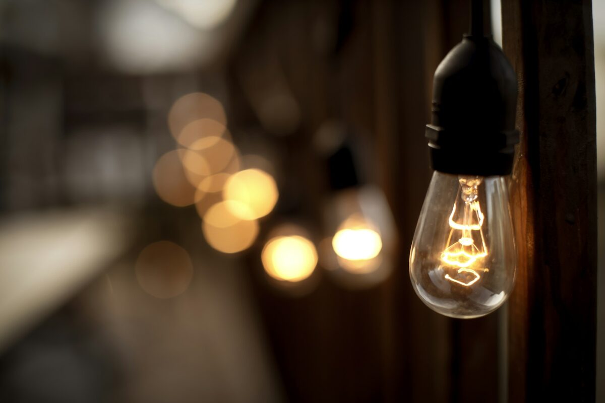 A clear filament bulb glowing warm amber from a black socket, the rest of its string blurred behind it