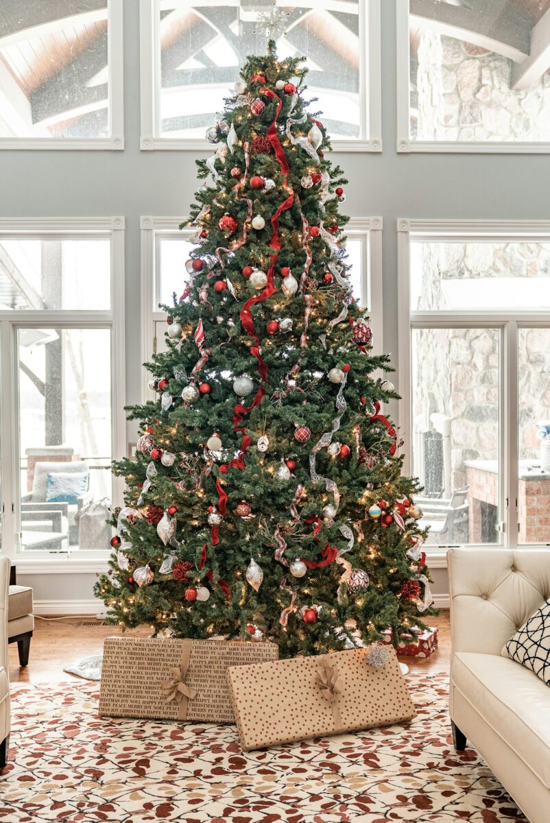A tall Christmas tree with red ribbon cascading loosely from top to bottom in a bright room with big windows