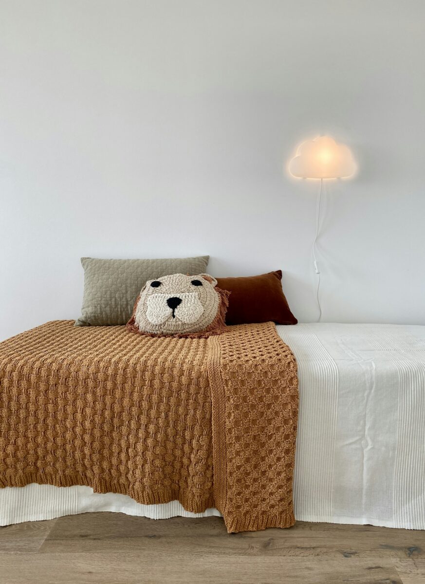 A caramel waffle-knit throw over a white bed with linen pillows and a glowing cloud wall light