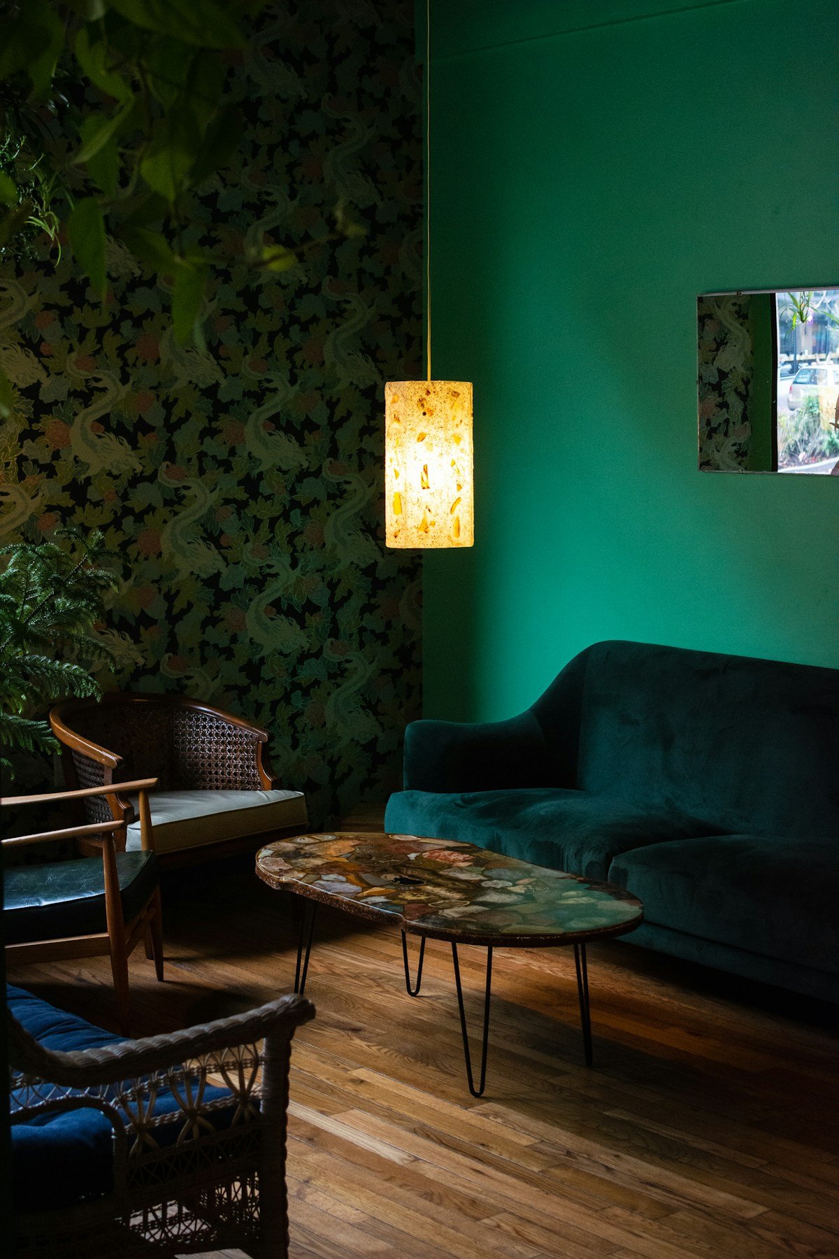 Large-scale leaf print wallpaper on a dark ground behind rattan armchairs, with a green velvet sofa and a wood floor