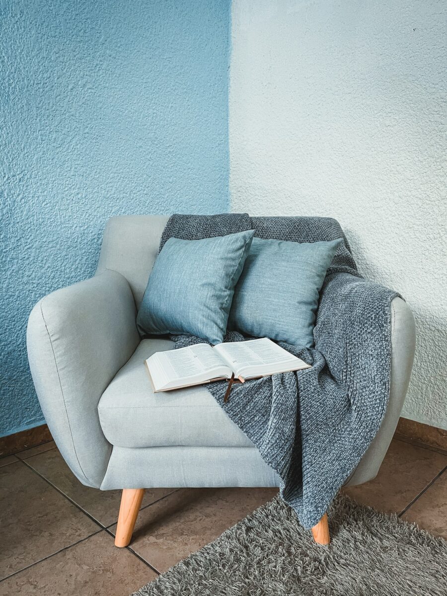 Light gray-blue mid-century armchair in a corner where one wall looks mid sky blue and the textured other wall looks paler and icier, with dusty blue linen pillows, a gray-blue knit throw, an open book and a gray shag rug