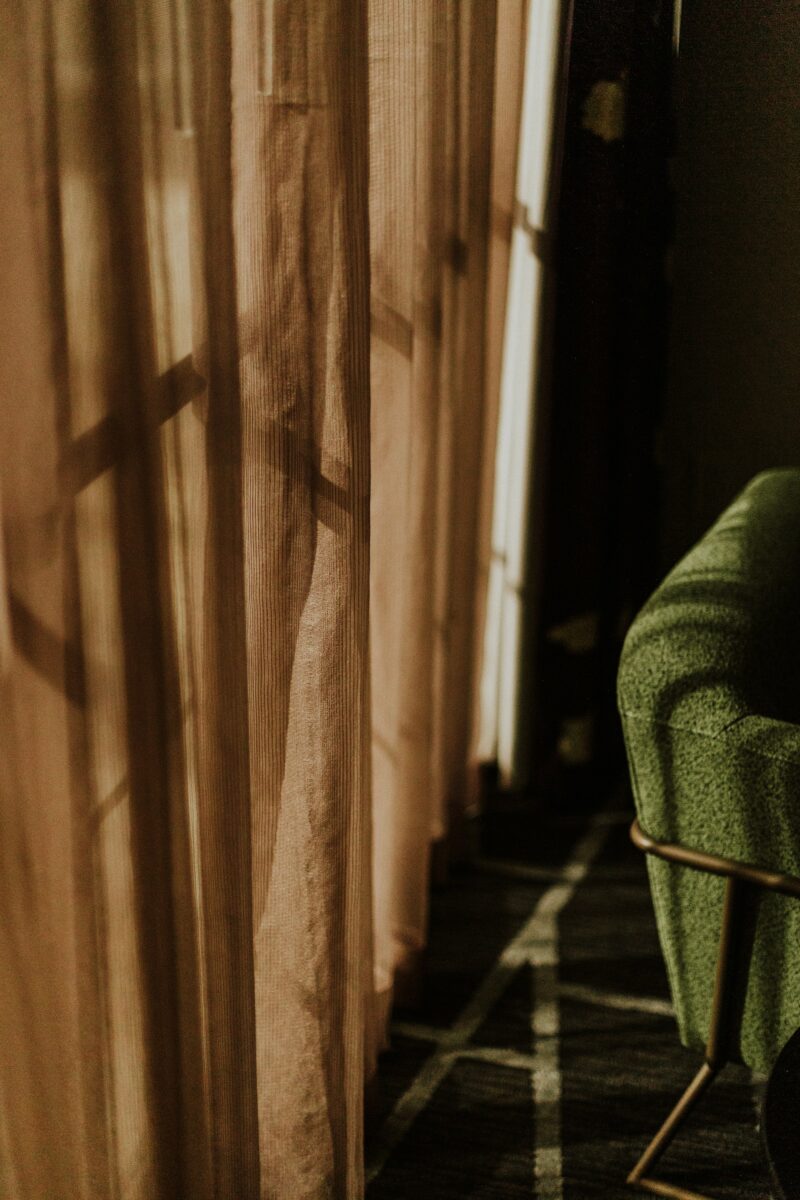 Sheer curtains glowing amber in late sun with window frame shadows, beside the arm of an olive armchair with a gold-toned metal leg on a dark carpet
