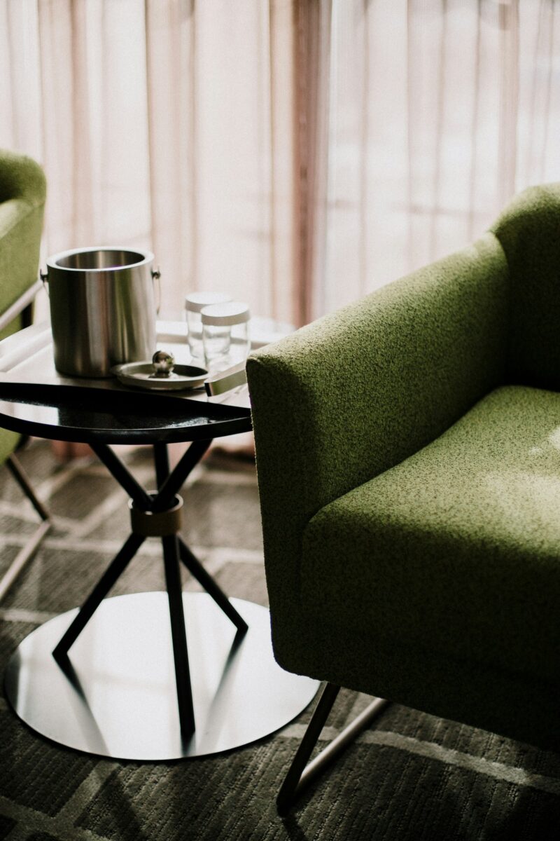 Two olive green nubby armchairs on metal sled legs beside a black side table with a steel disc base, holding an ice bucket and two glasses, in front of backlit blush pink sheer curtains