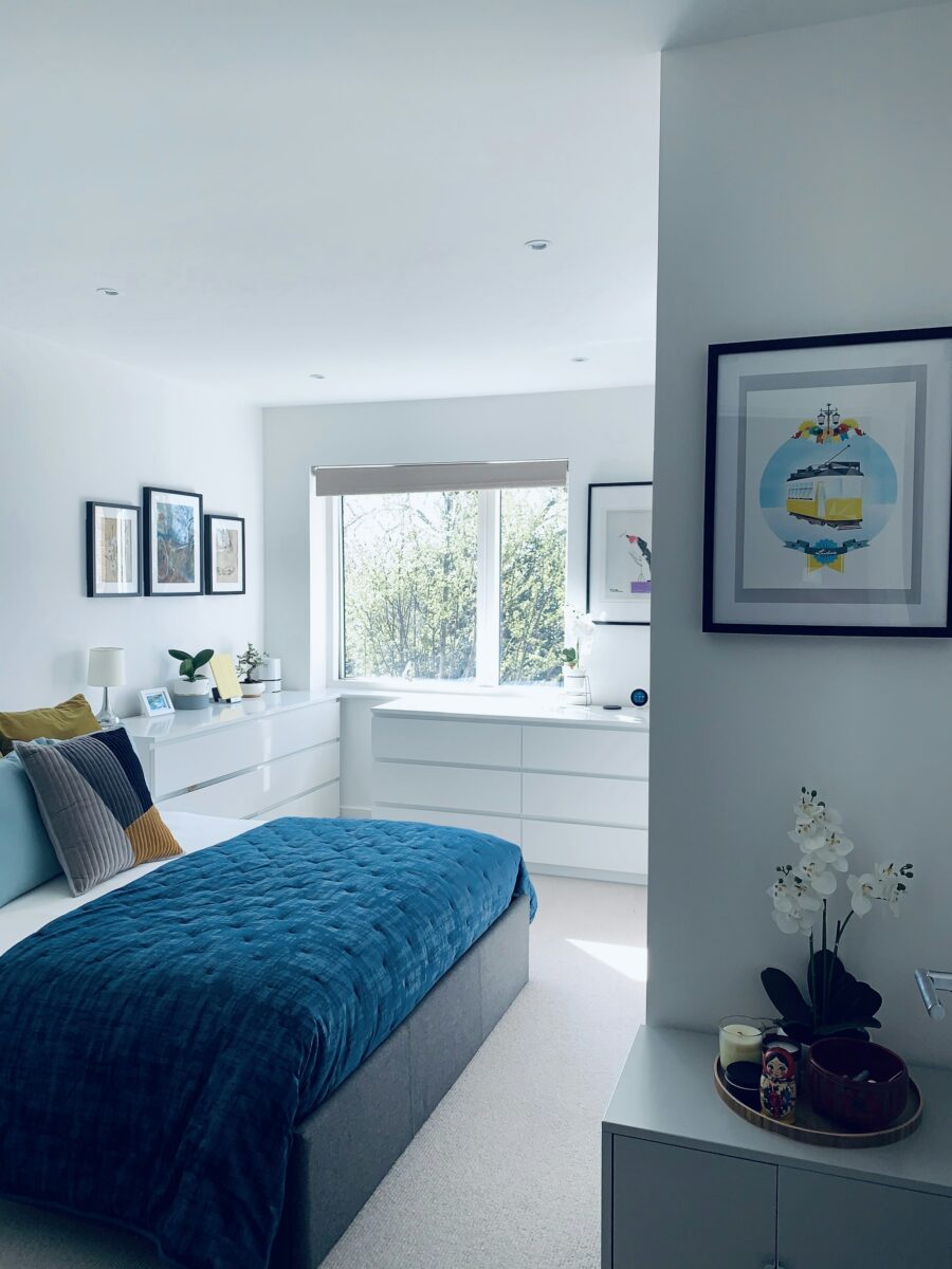 Bedroom with white walls and a gray upholstered bed made with a deep teal blue velvet quilt, a pale blue pillow and a mustard and gray pillow, white dressers under the window and black-framed prints including a tram print with a light blue circle