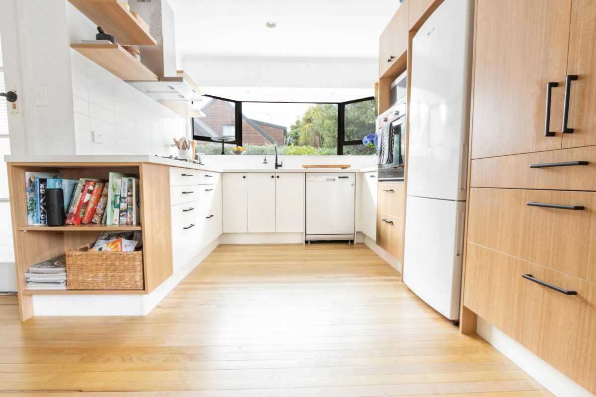 U-shaped kitchen with white lower cabinets and black bar pulls, tall light oak cabinets with long black pulls, an open oak cookbook shelf with a woven basket, black-framed windows over the sink and a honey wood floor