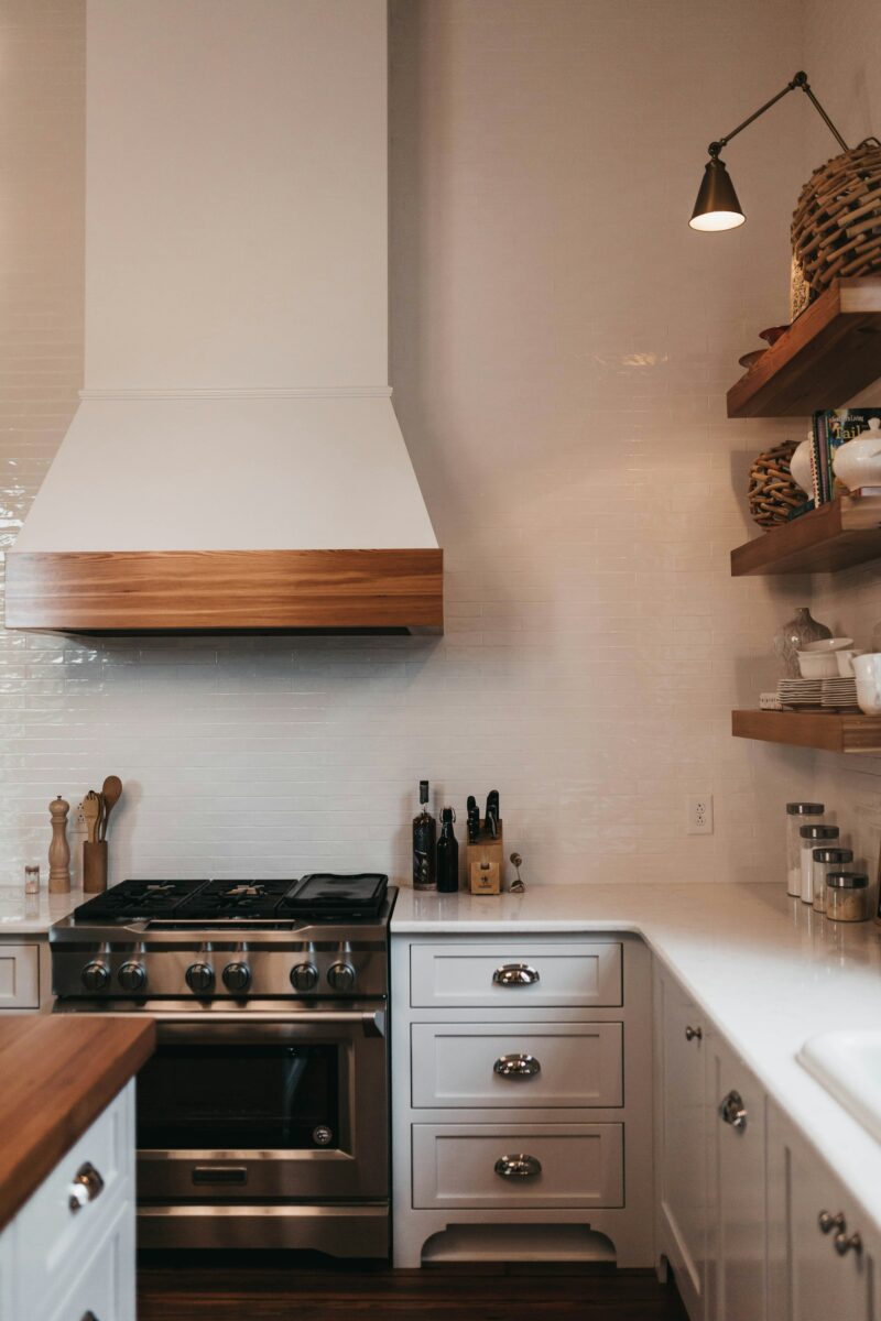 White plaster-look range hood with a wood band, small glossy white tile up to the ceiling, white shaker drawers with nickel cup pulls and a lit swing-arm wall sconce over floating wood shelves