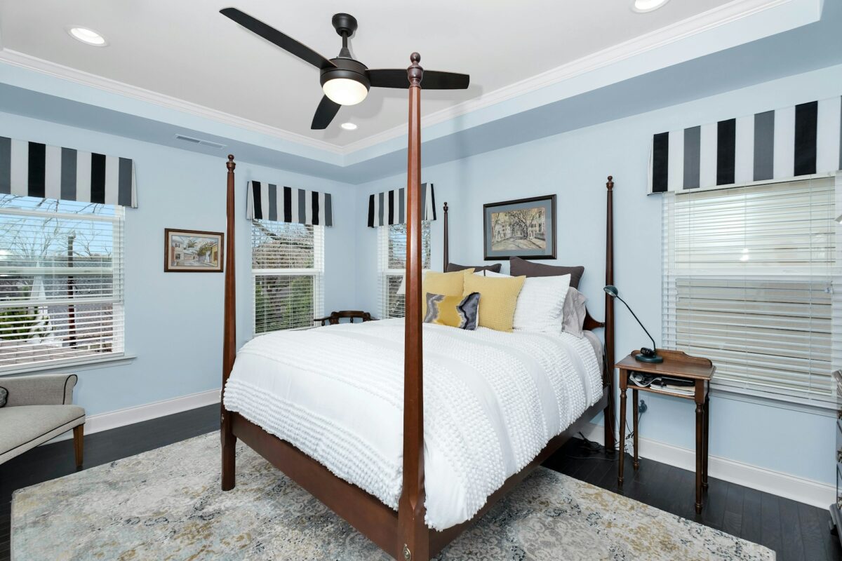 The same light blue bedroom seen from the window side, with windows on two walls under striped valances, a small wood nightstand with a black task lamp and the ceiling fan light on