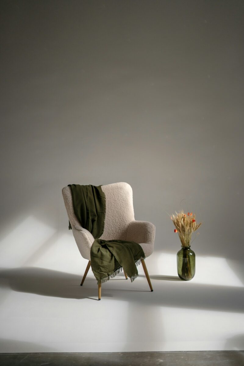 Cream boucle armchair with tapered wood legs, an olive fringed throw over the arm and an olive glass vase with dried wheat and small orange-red flowers in warm raking sunlight