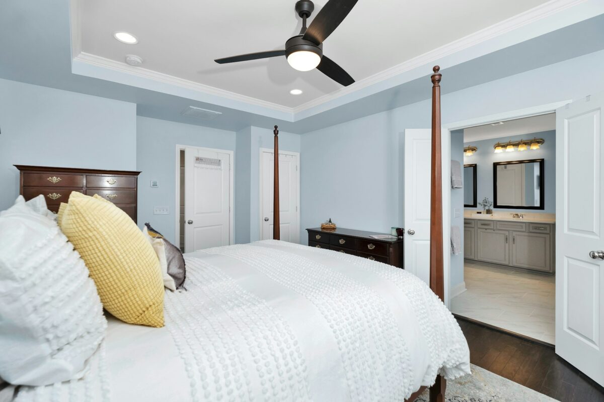 The light blue bedroom looking toward an en-suite bathroom painted the same blue, with a gray vanity, black-framed mirrors and a brass vanity light, dark wood chests with brass pulls and a white bedspread with a yellow pillow