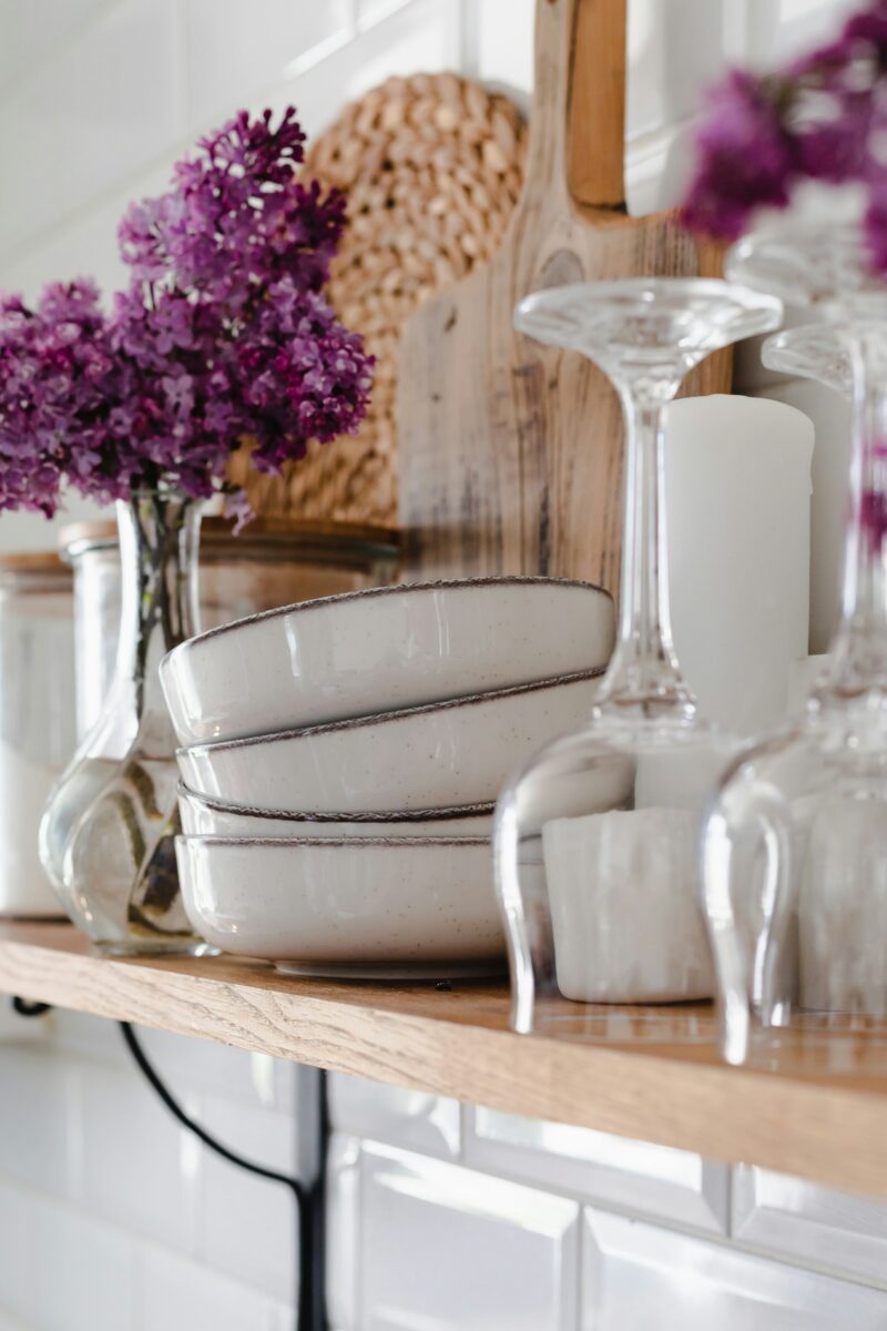 Oak shelf on black brackets with speckled stoneware bowls with brown rims, upturned wine glasses, a white pillar candle, lilac in a glass vase, a worn wooden board and a rattan trivet against white beveled subway tile