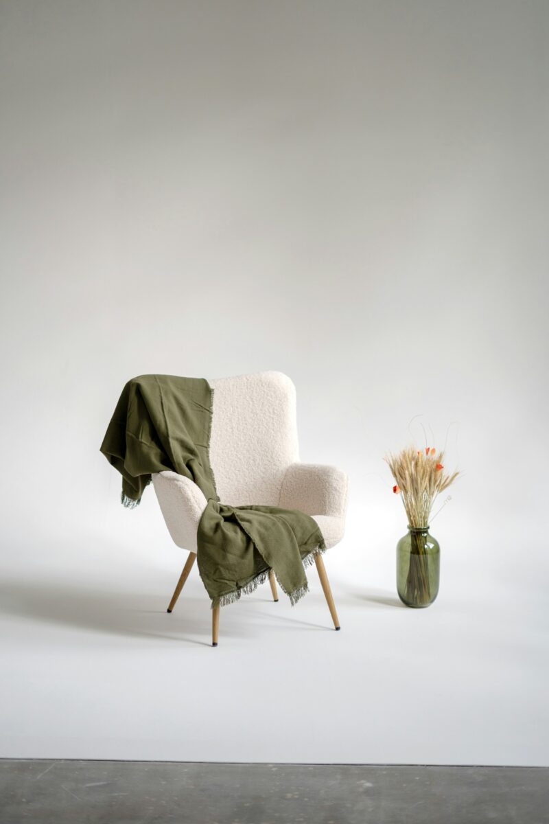 The same cream boucle chair, olive fringed throw and olive vase with dried wheat in flat, even light against a white-looking wall