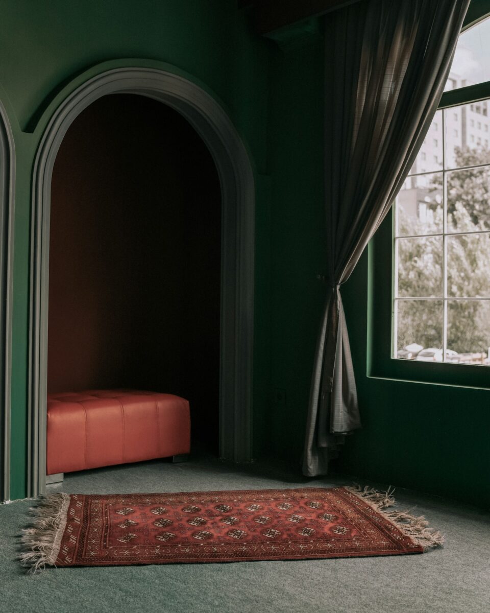Red rug with rows of octagon medallions on a gray-green carpet in a dark green room with a coral tufted leather bench in an arched alcove