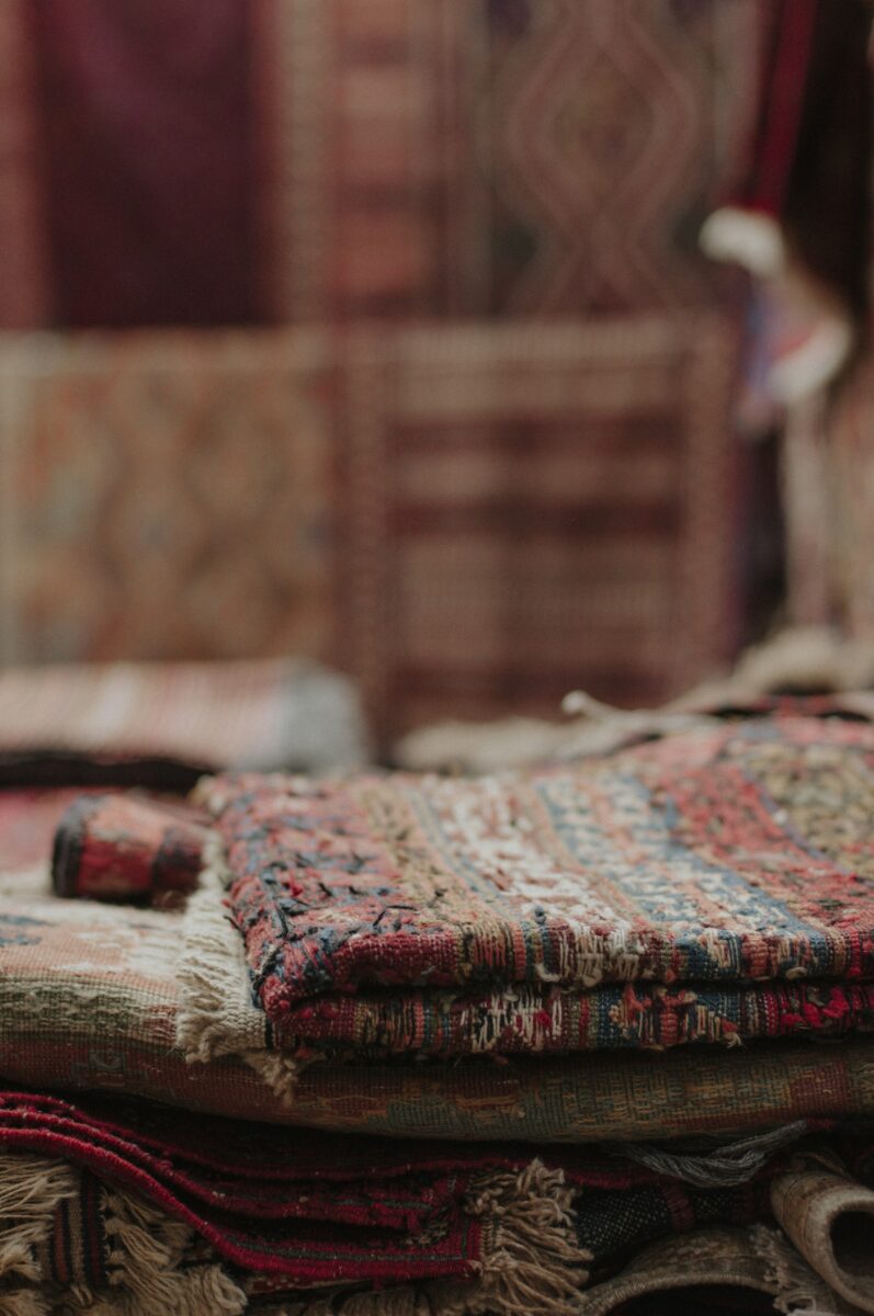 Close-up of a stack of folded, well-worn rugs and flatweaves with loose yarn ends showing, more rugs hanging behind