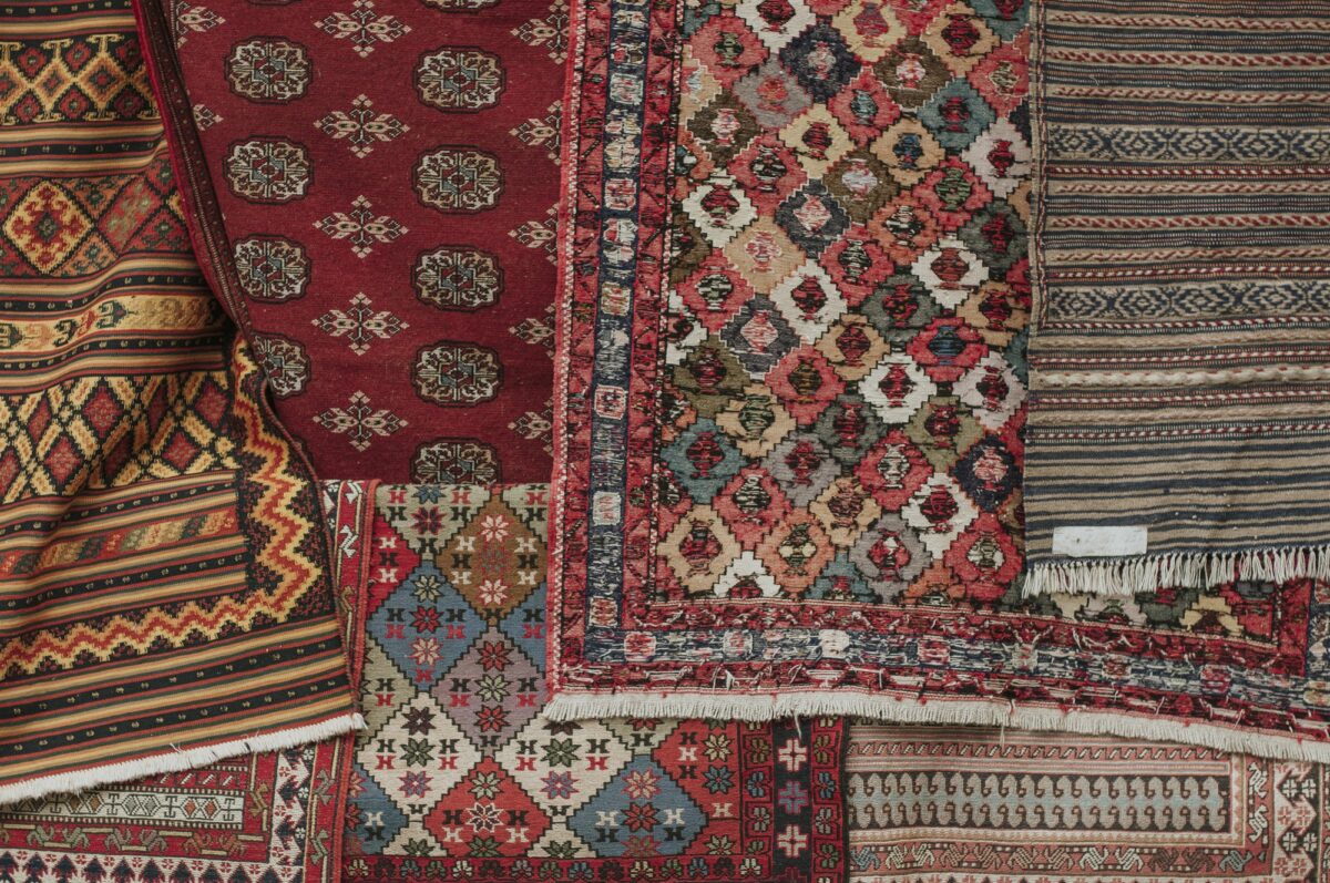 Overhead view of overlapping rugs, a red rug with rows of octagon guls, a multicolor diamond lattice rug and striped kilims with a white tag