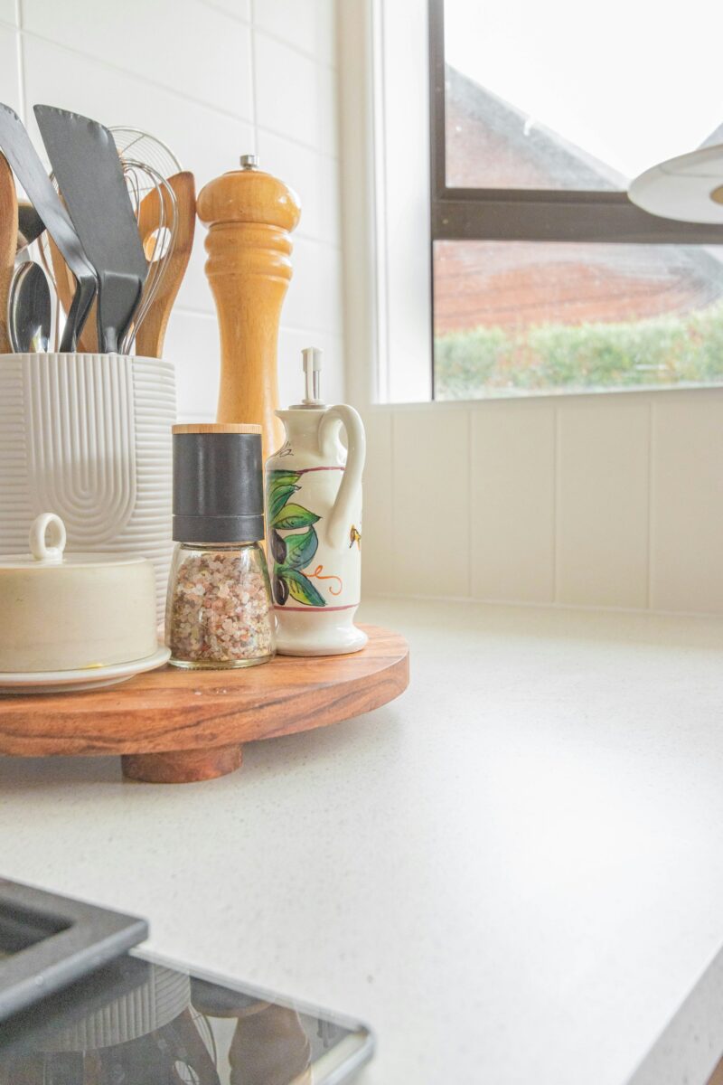 Wooden turntable on a white speckled counter holding a ribbed white utensil crock, a wood pepper mill, a salt grinder, a hand-painted olive oil cruet and a butter dish
