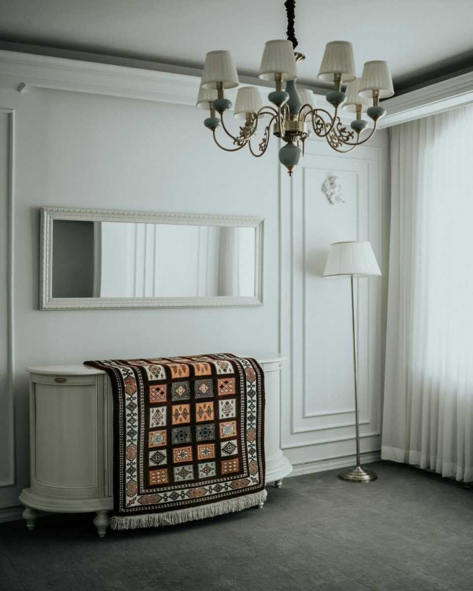 Geometric rug with a grid of square diamond panels draped over a cream oval sideboard in a white paneled room with a chandelier and a brass floor lamp