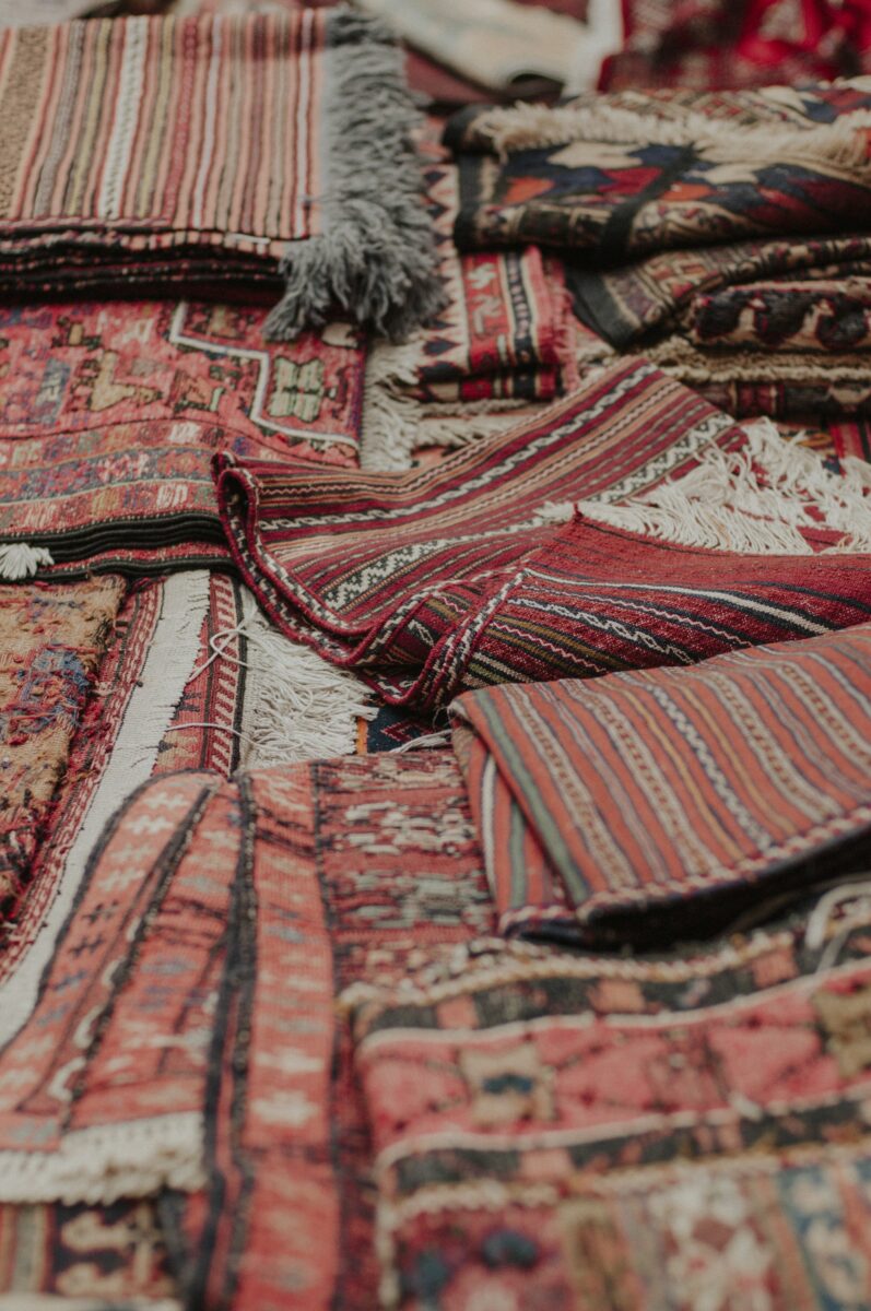 Piles of folded red striped flatweaves and pile rugs with fringe, the reds faded to soft rust and pink tones