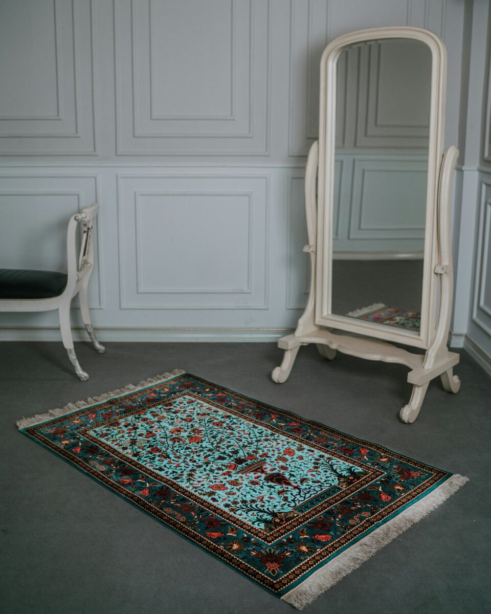 Small turquoise Persian-style rug with tree and bird motifs on gray carpet, beside a cream floor mirror and a white bench with a dark green velvet seat