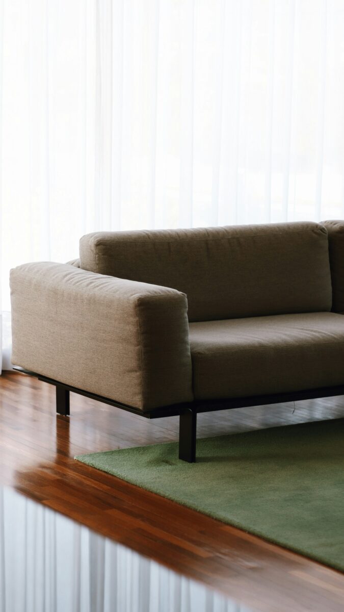 Low taupe sofa on a black frame over a solid olive green rug on a glossy red-brown wood floor with sheer white curtains