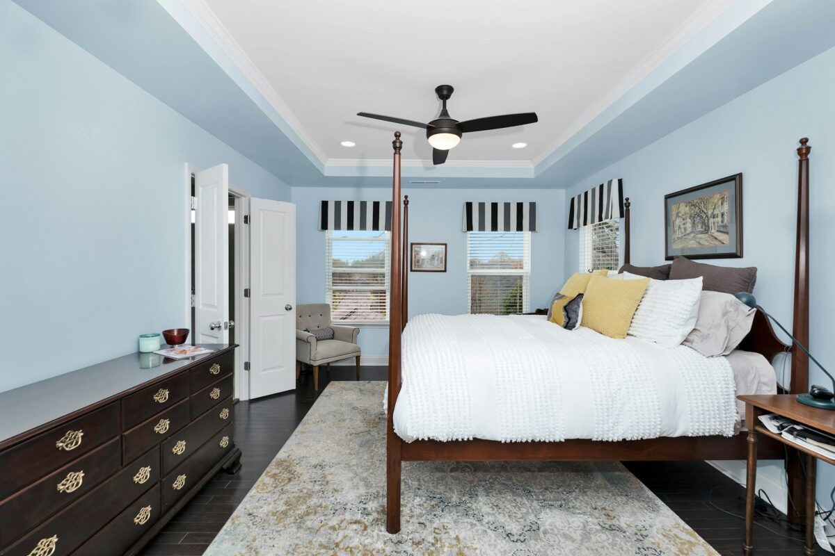Bedroom with powder blue walls, a white tray ceiling, a dark cherry four-poster bed with a white chenille bedspread and yellow and gray pillows, a long dark dresser with brass pulls and striped valances