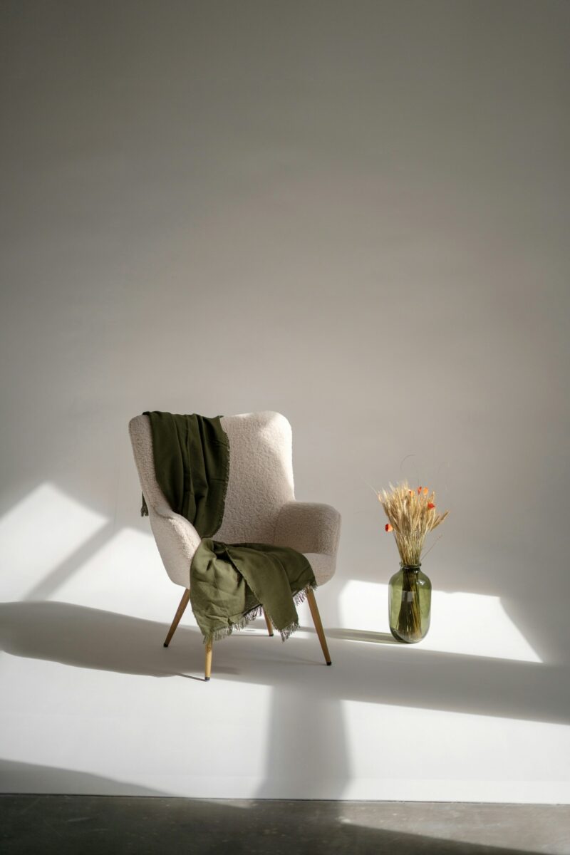 The same cream chair with the olive throw and vase, with window-shaped patches of sunlight falling across the chair and wall