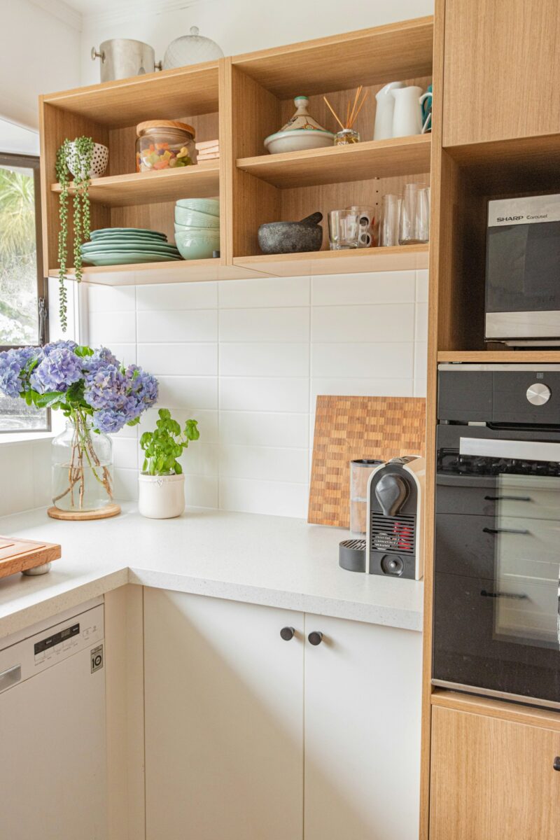 Kitchen corner with light oak open box shelves holding sage green plates and bowls, white flat-panel cabinets with small black knobs, a checkered end-grain cutting board, basil and blue hydrangeas in a glass vase