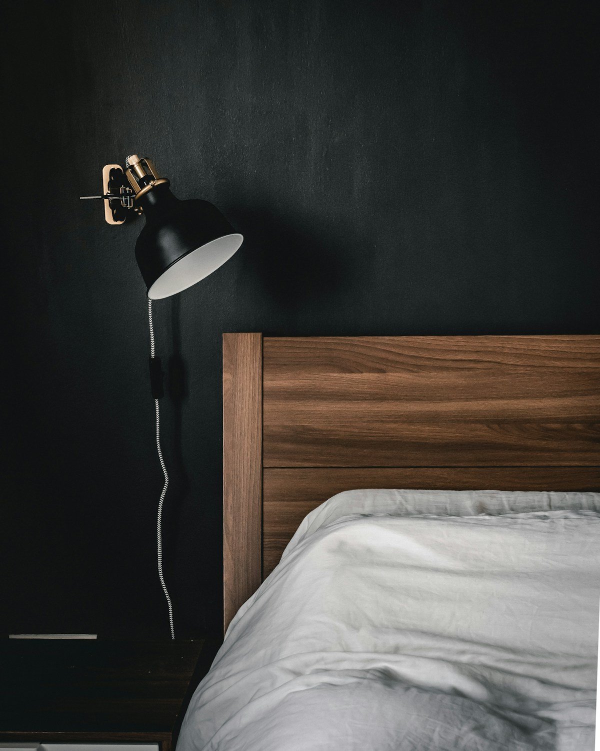 Walnut headboard and a black swing-arm wall lamp against a near-black bedroom wall